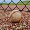 A baseball sits on the ground by a fence.