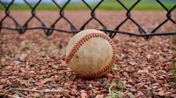 A baseball sits on the ground by a fence.